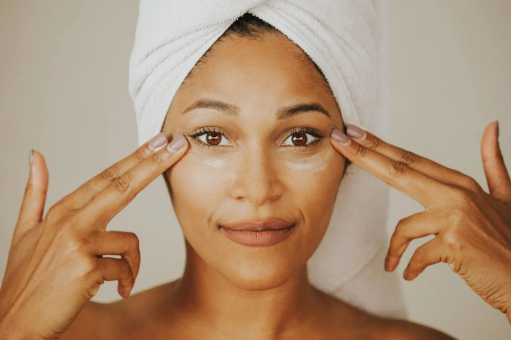 Uma mulher com uma toalha branca enrolada na cabeça aplica suavemente um creme na área abaixo dos olhos, usando os dedos de forma cuidadosa. Com uma expressão serena, ela demonstra um momento de autocuidado como parte de sua rotina de higiene pessoal. A imagem destaca a importância de cuidar da pele, transmitindo tranquilidade e bem-estar, além de reforçar a relevância dos cuidados com a higiene pessoal na rotina diária de beleza.