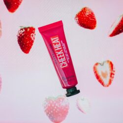 Um tubo de blush em gel da Maybelline, chamado "Cheek Heat", de cor rosa vibrante, flutua em frente a um fundo decorado com imagens de morangos inteiros e cortados ao meio. O produto é destacado ao centro, contrastando com o fundo rosado e a textura dos morangos.