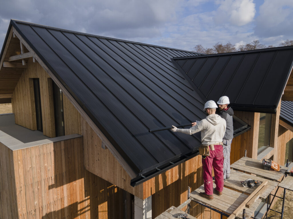 Dois trabalhadores da construção civil, utilizando capacetes brancos, estão instalando telhas pretas em um telhado de uma casa moderna com paredes de madeira e elementos pré moldados.
