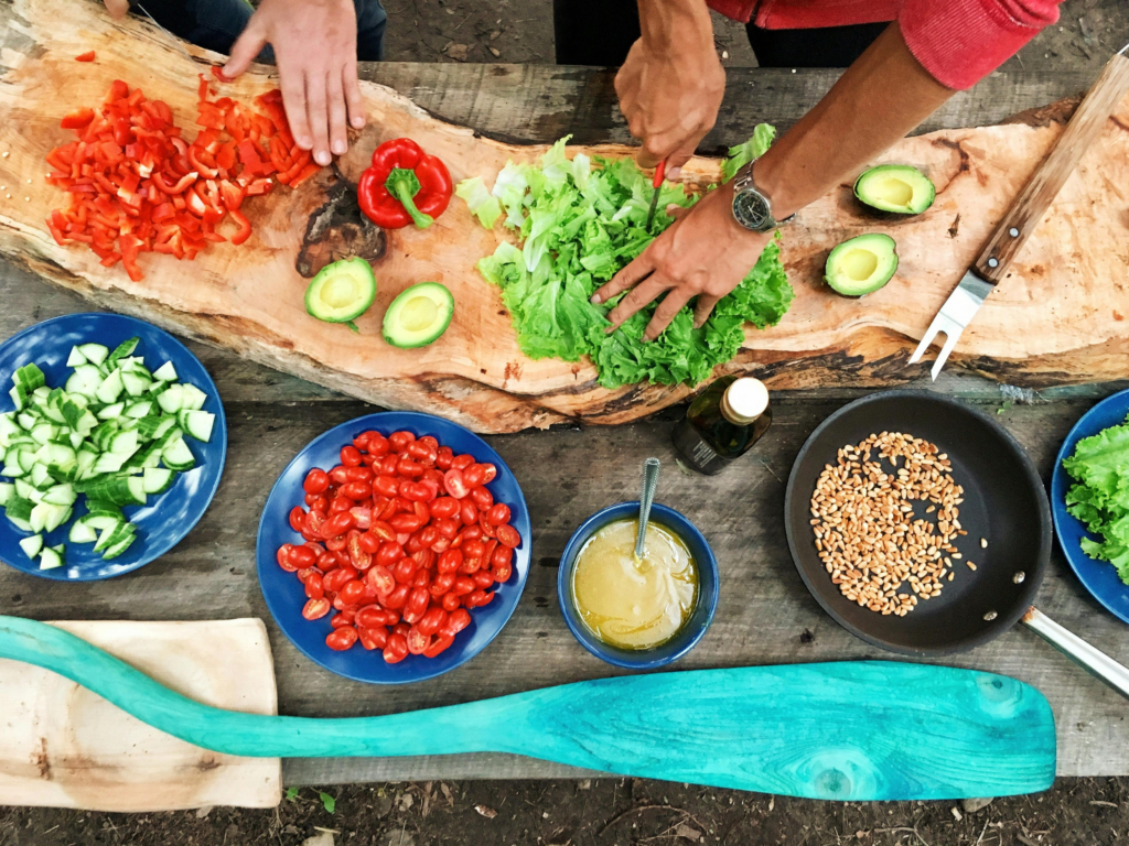 Visão de cima de uma mesa de madeira com pimentões picados, alface sendo cortada, metades de abacate, tomates cereja, pepino em um prato, sementes em uma frigideira e molho. A manipulação de múltiplos alimentos crus ressalta a necessidade de prevenir a contaminação cruzada.