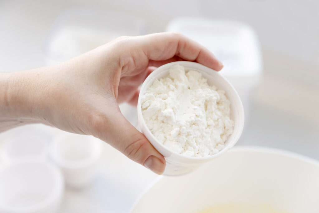 Uma mão segura uma pequena tigela branca cheia de um pó branco, que pode ser creatina.