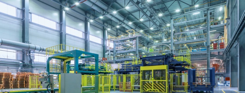 Interior de uma fábrica moderna com layout industrial eficiente, equipada com máquinas automatizadas, estruturas metálicas, esteiras e áreas de circulação delimitadas por grades de segurança. O ambiente é amplo, bem iluminado e organizado para otimizar o fluxo produtivo e a operação dos equipamentos.
